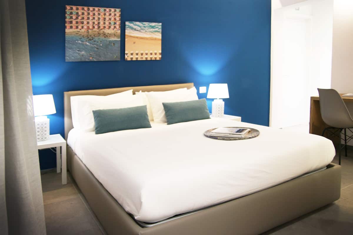 Una cama bien hecha con almohadas blancas y verdes, una mesita de noche con lámpara y dos cuadros en la pared.