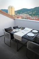 Un balcón con una mesa dispuesta para dos, con vista a un paisaje urbano de edificios y colinas.