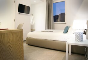 Una habitación de hotel moderna con una cama grande, un televisor de pantalla plana montado en la pared, una mesita de noche con lámpara y vista a la ciudad al anochecer a través de la ventana.