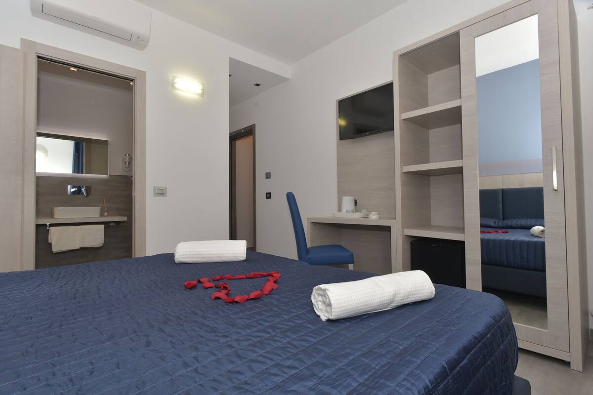 A hotel room with a large bed, a desk, a chair, a TV, and a wardrobe.