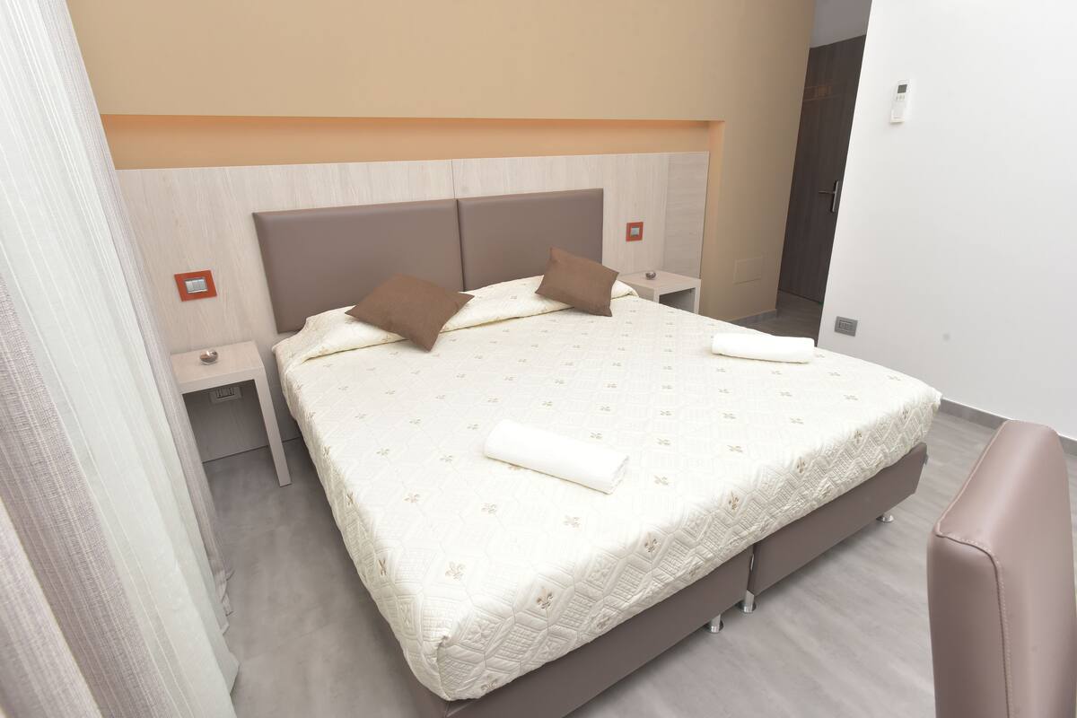 A neatly made bed with a patterned bedspread, two pillows, and a rolled towel. There are two bedside tables with lamps. The room has a light-colored wooden headboard and a plain white wall.