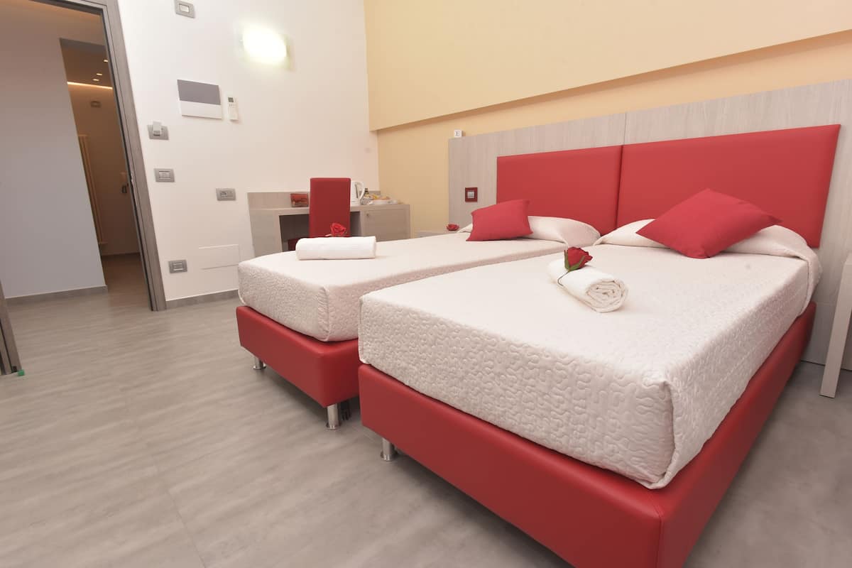 A hotel room with two beds, each with red headboards and white bedding. There is a small desk with a chair and a red flower vase.