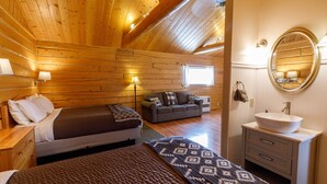 A cozy log cabin room with a bed, a sofa, a round mirror, and a bathroom with a sink. - Sheep Mountain Lodge (Sutton)