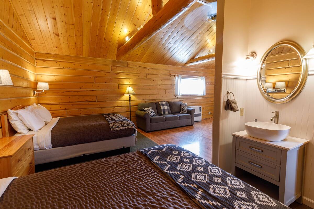 A cozy log cabin room with a bed, a sofa, a round mirror, and a bathroom with a sink.