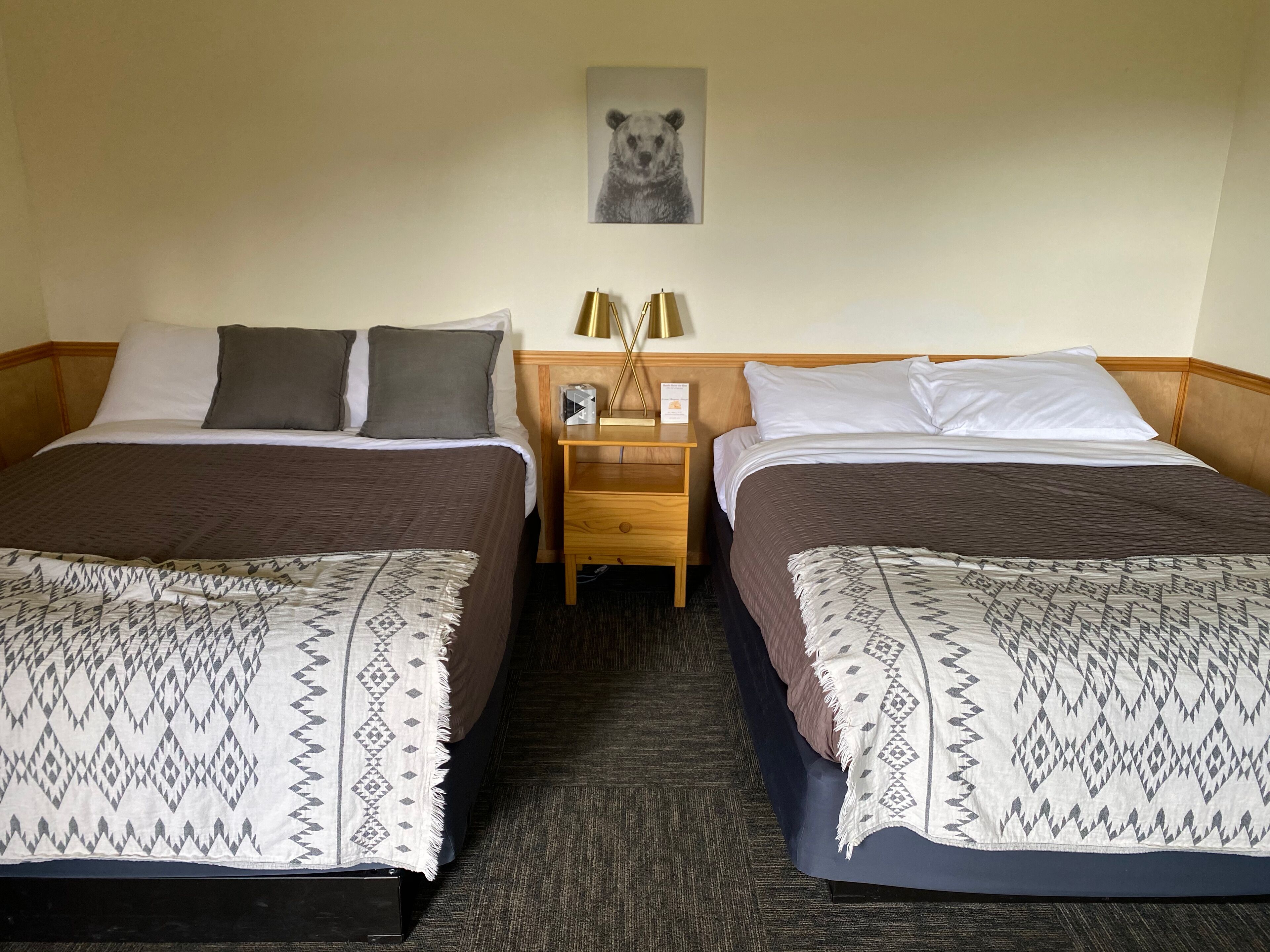 Ein Hotelzimmer mit zwei Betten, jeweils mit gemusterten Bettdecken und Kissen. Ein Nachttisch mit Lampe und ein gerahmtes Bild an der Wand über den Betten.