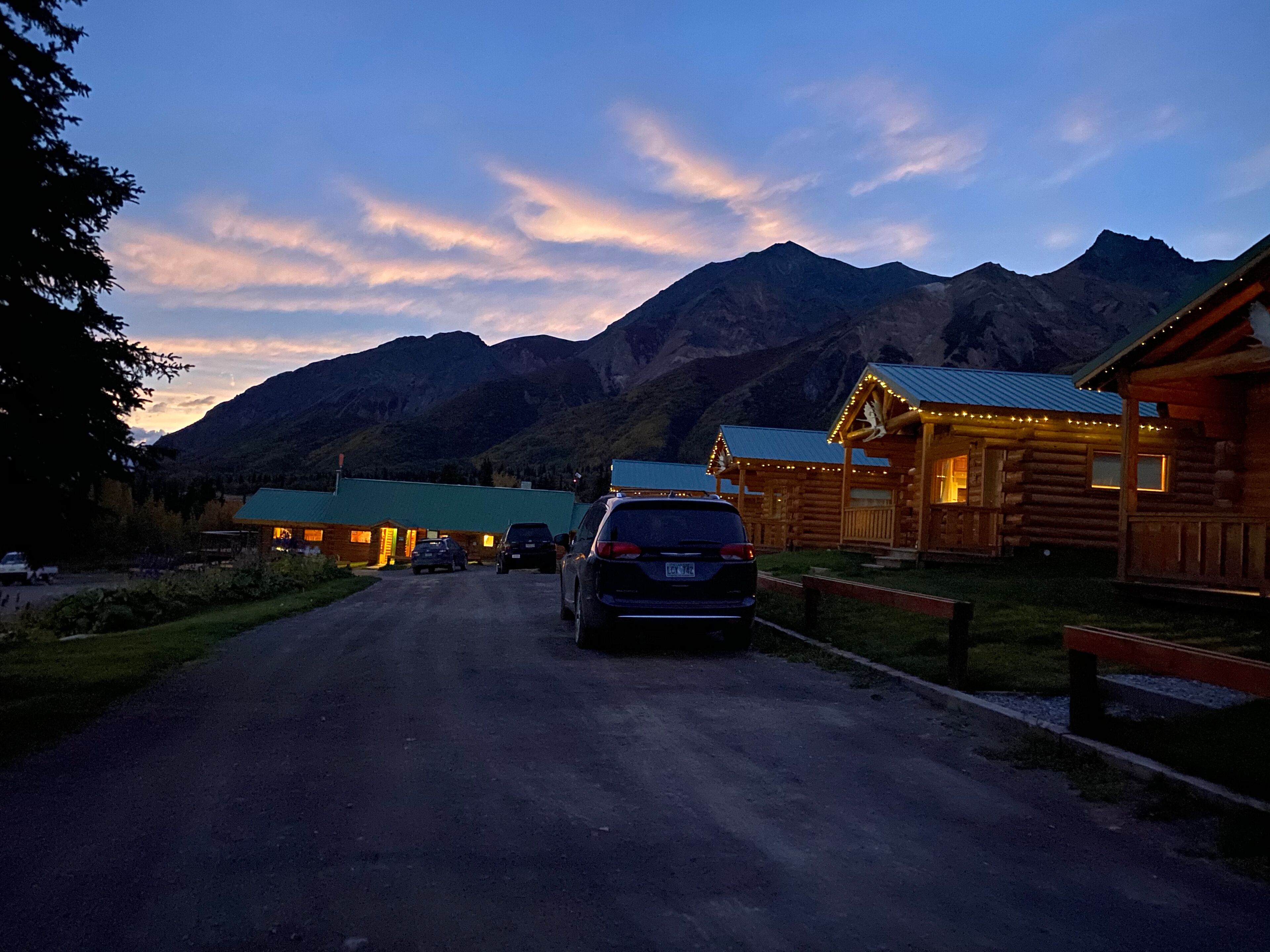 A log cabin-style building with a green roof, a car parked in front, and mountains in the background.