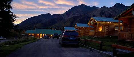 A log cabin-style building with a green roof, a car parked in front, and mountains in the background.