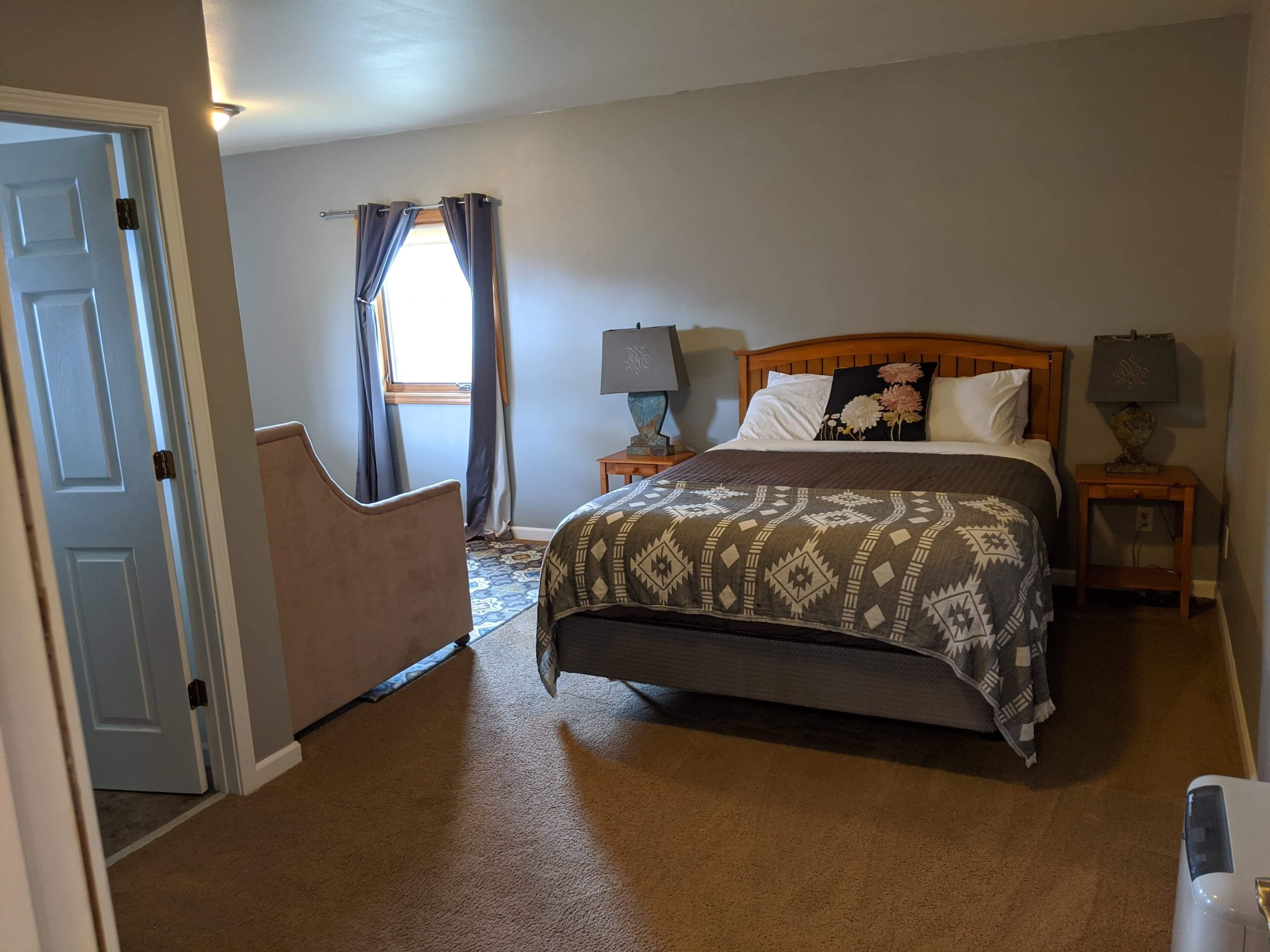 A bedroom with a bed, two bedside tables with lamps, a window with curtains, and a door.