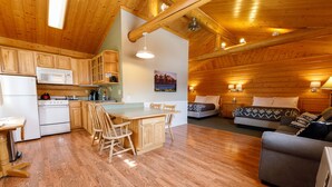 A cozy cabin-style room with wooden walls and floors, a kitchenette, a dining area with a table and chairs, and a comfortable seating area with a sofa and a bed. - Sheep Mountain Lodge (Sutton)