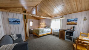A cozy cabin-style room with a bed, a small kitchenette, a dining area, and a living space with a sofa and a window overlooking the interior. - Sheep Mountain Lodge (Sutton)