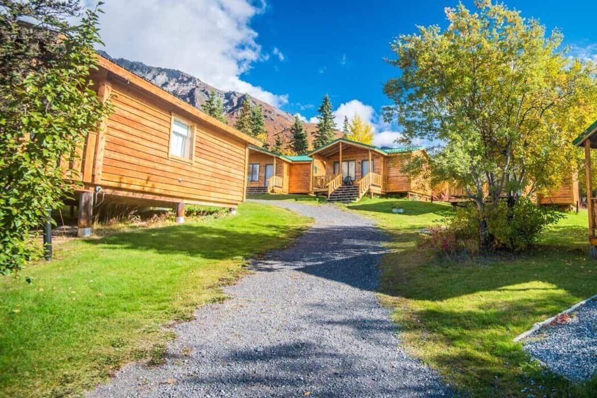 A row of wooden cabins with a gravel path leading through a grassy area, surrounded by trees and mountains in the background.