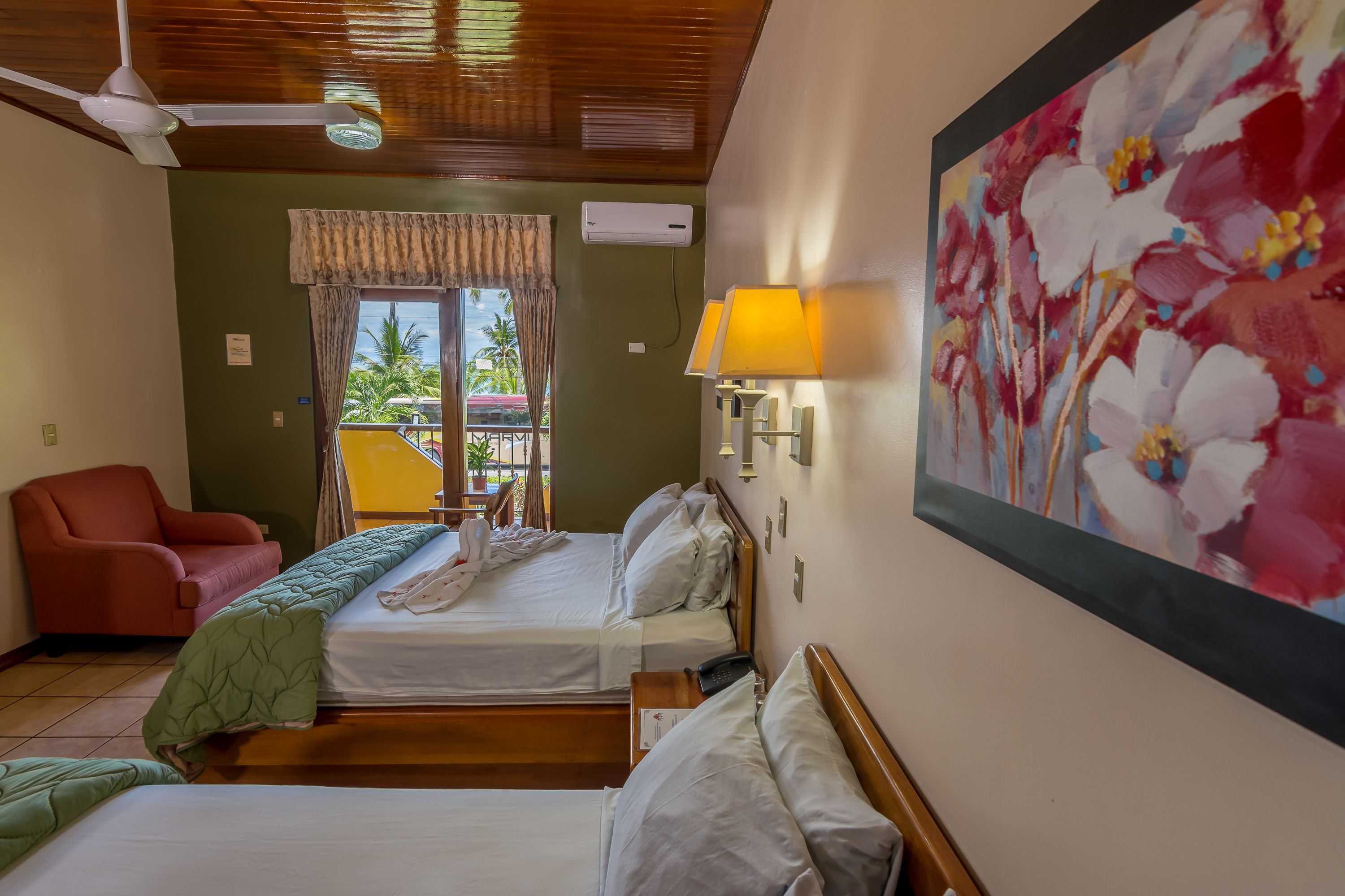 A hotel room with two beds, a red armchair, a painting, and a balcony with a view of palm trees.