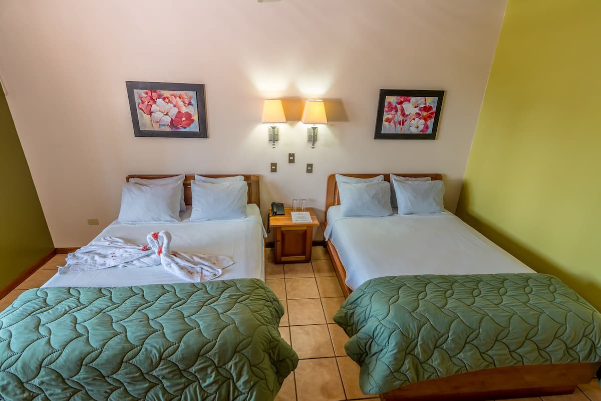 A hotel room with two single beds, each with a green quilted bedspread and white pillows. There are two wall-mounted lamps between the beds, and two framed floral paintings above them.