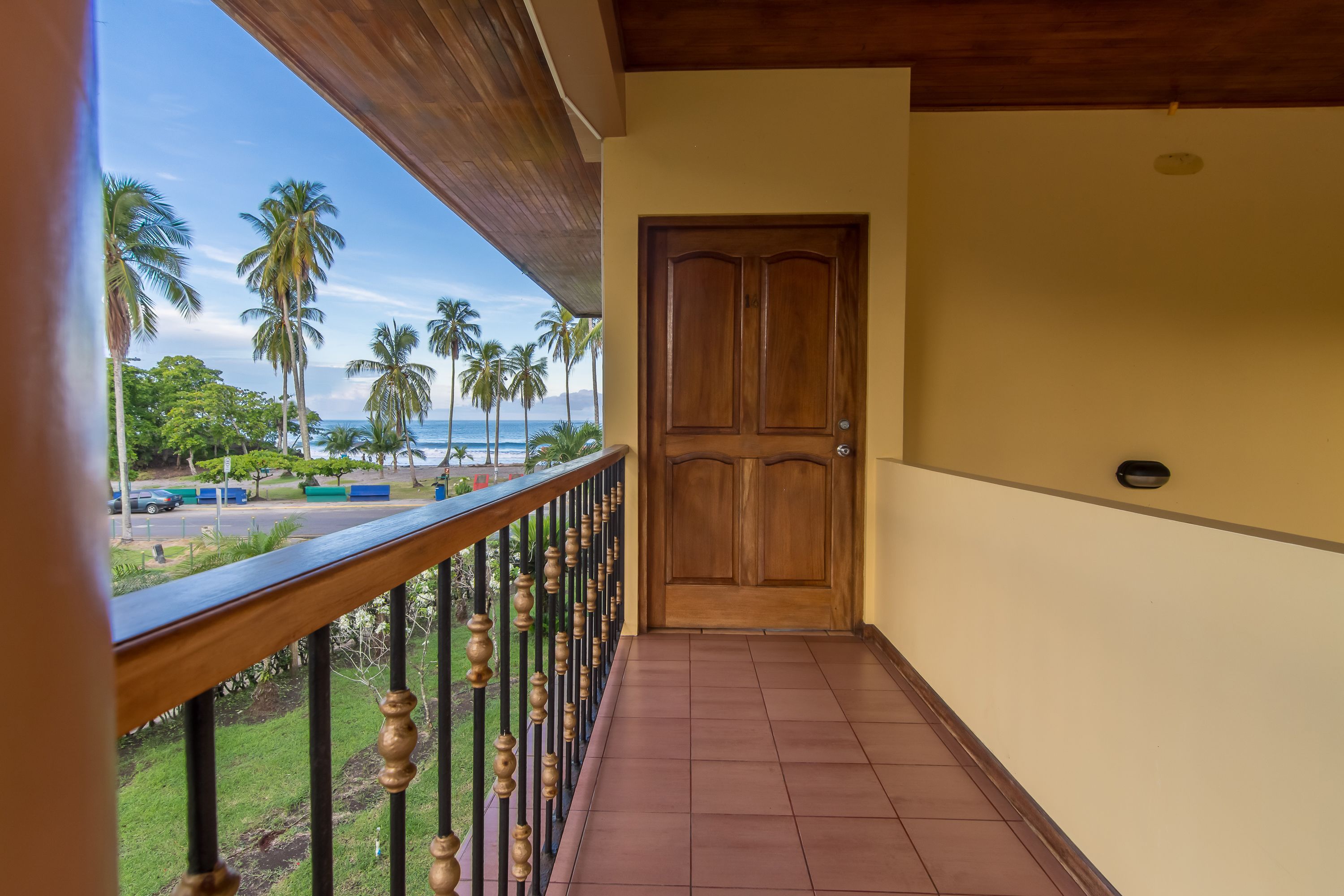 A balcony with a wooden railing, a closed wooden door, and a view of palm trees and a road.