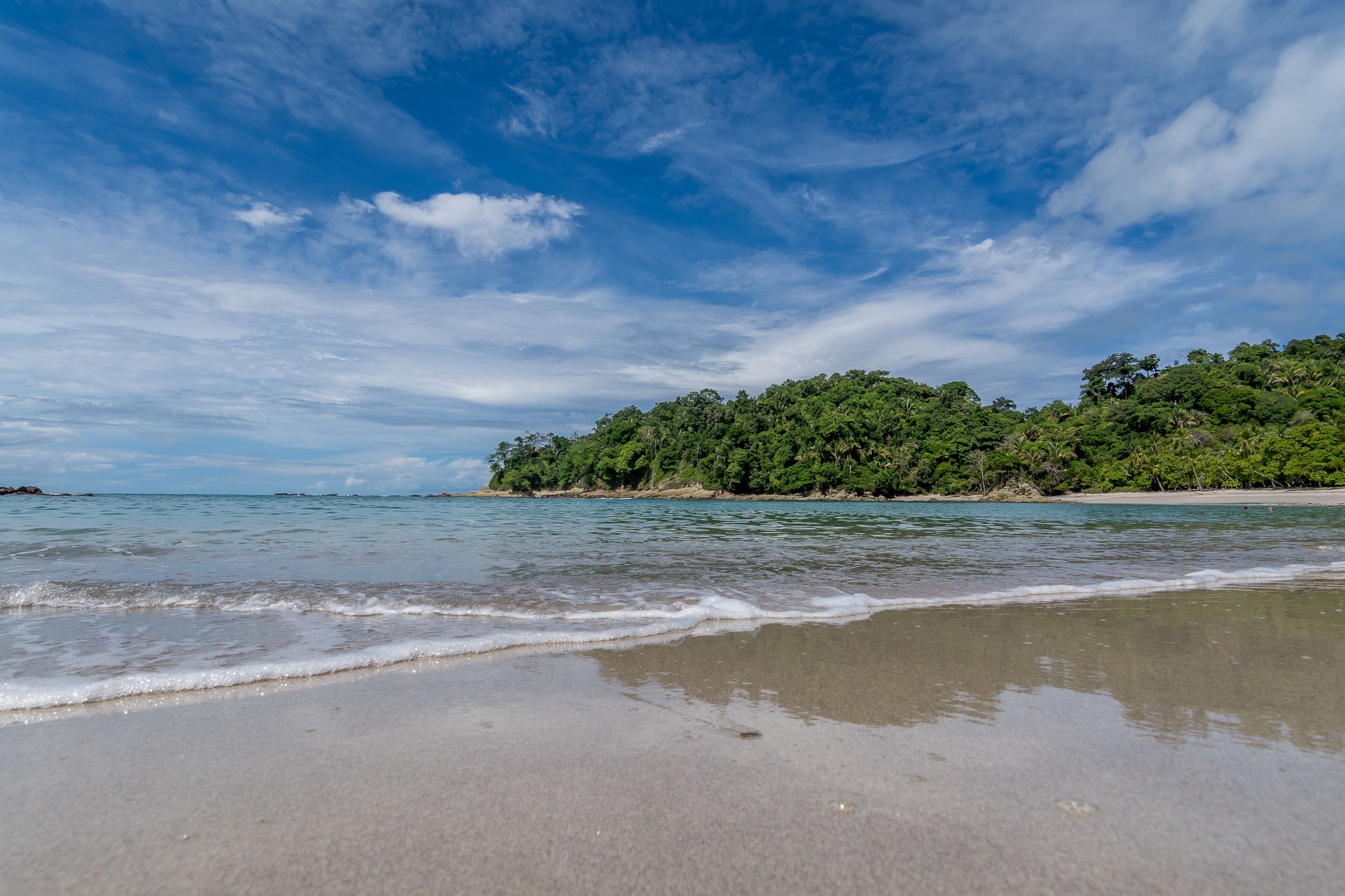 Una playa con agua azul cristalina, orilla de arena y una colina boscosa al fondo.