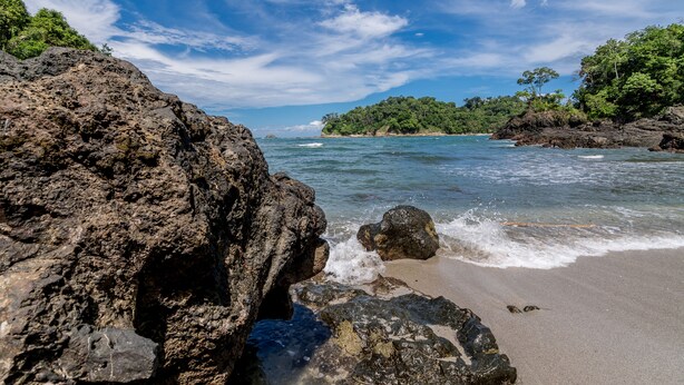 A rocky shoreline with clear blue water, a sandy beach, and lush greenery in the background. - Hotel Manuel Antonio (Quepos)