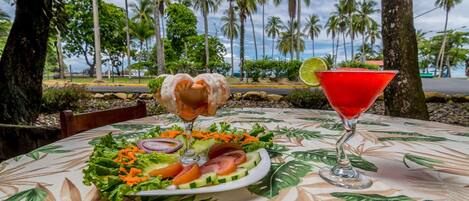 A table with a tropical leaf-patterned tablecloth, a salad, and two drinks, set against a backdrop of palm trees and a road.