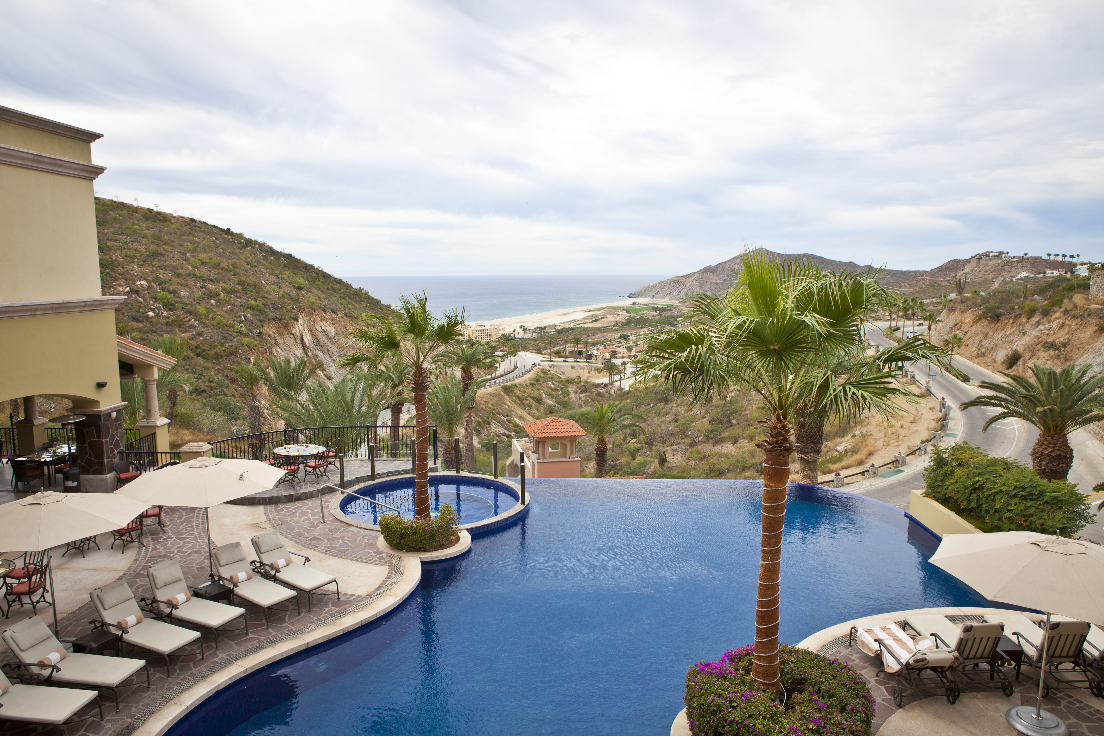 Piscina de resort con sillas de descanso y palmeras, con vistas a una playa y un paisaje montañoso.