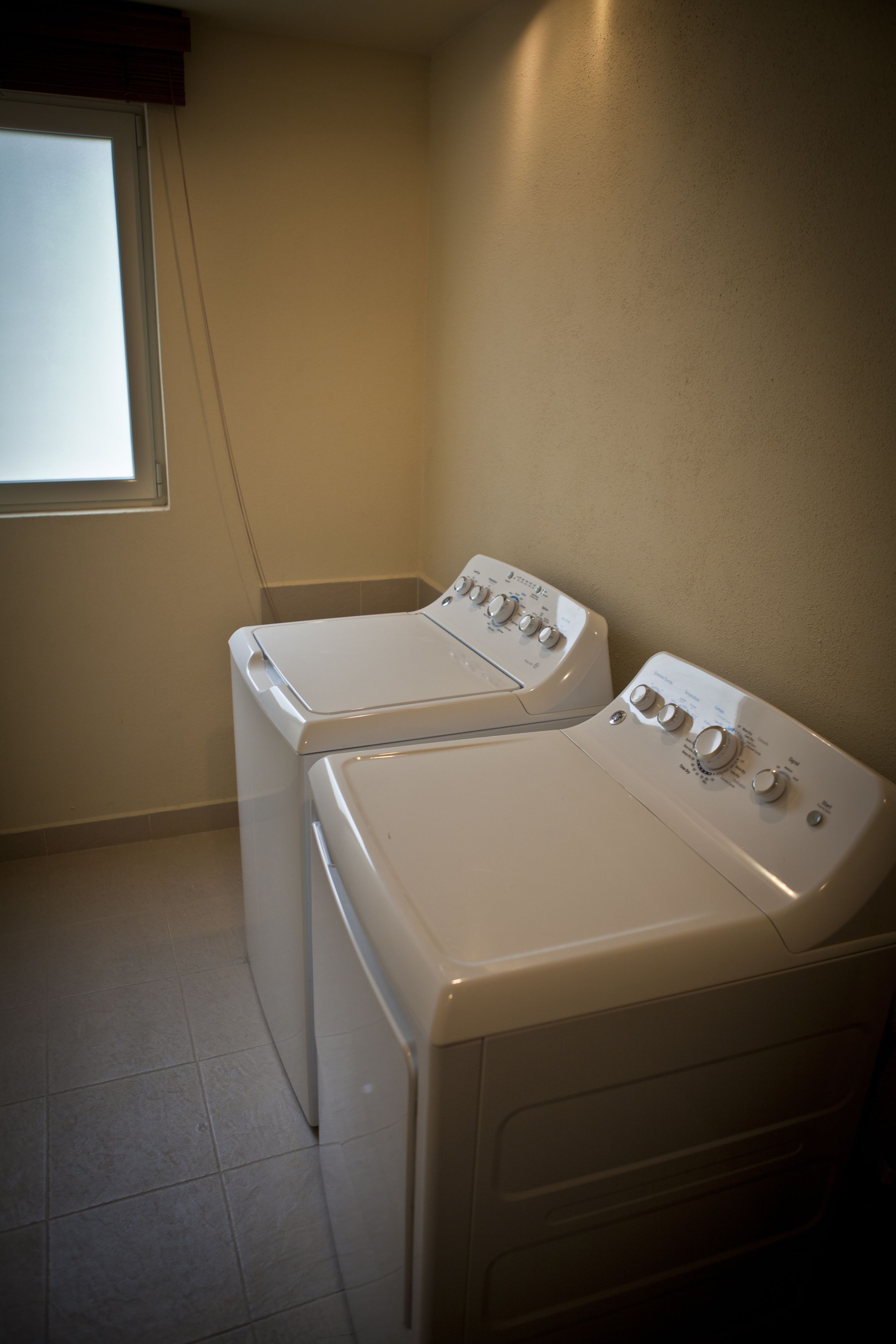 A laundry room with two stacked front-loading washing machines and a window with blinds.