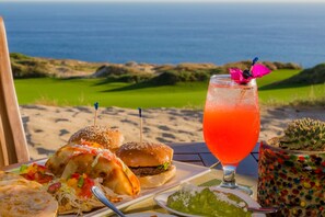 Un repas composé d’un hamburger et de tacos, un verre avec une garniture, et un cactus dans un pot coloré, le tout sur une table offrant une vue sur l’océan et les collines verdoyantes.