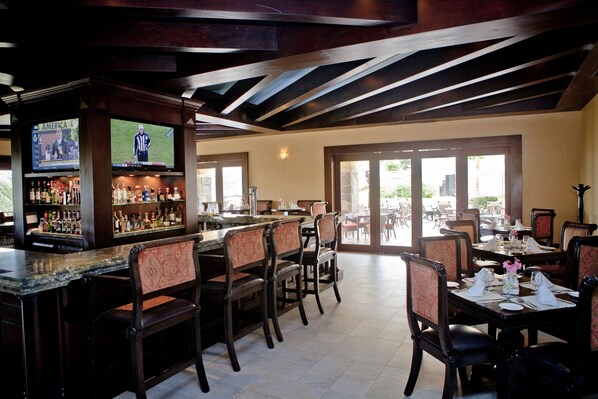 A bar area with a television displaying a sports game, a bar counter with various bottles, and several dining tables with chairs. - Quivira Los Cabos Condos & Homes - Vacation Rentals (Cabo San Lucas)