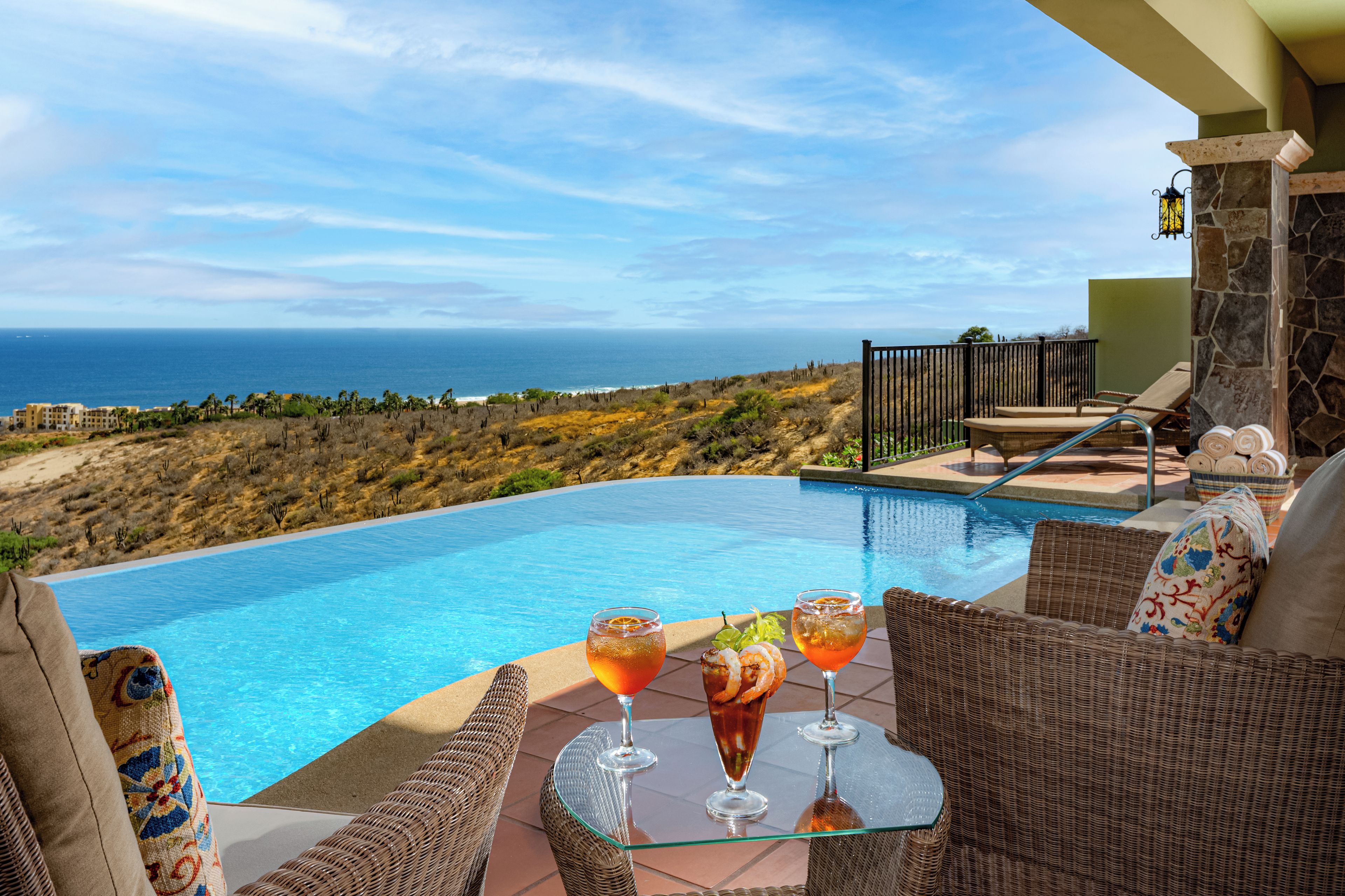 A poolside area with a view of the ocean, wicker furniture, and two drinks on a glass table.