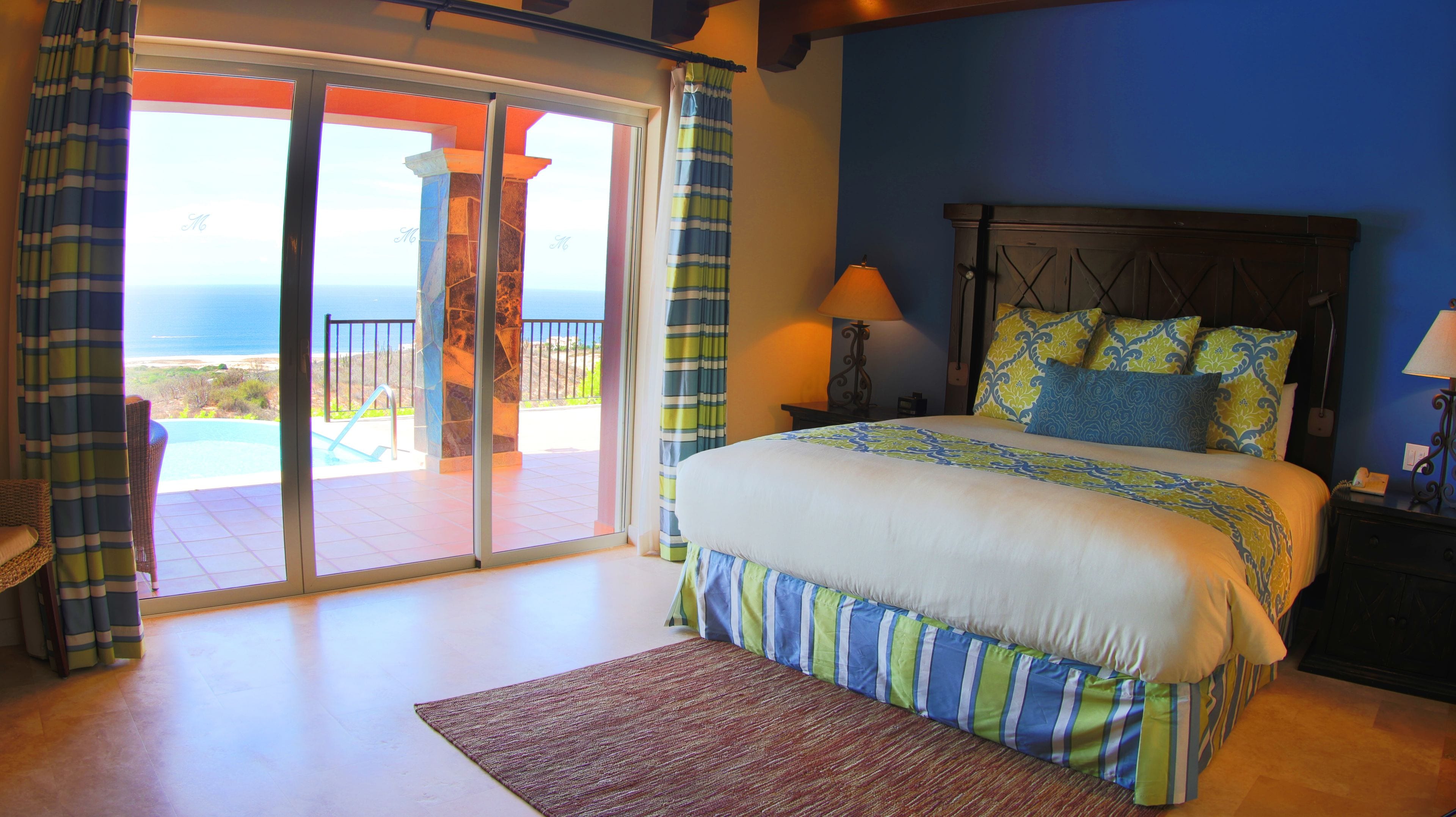 Un dormitorio con cama, mesita de noche, lámpara y vista al océano a través de una puerta corrediza.