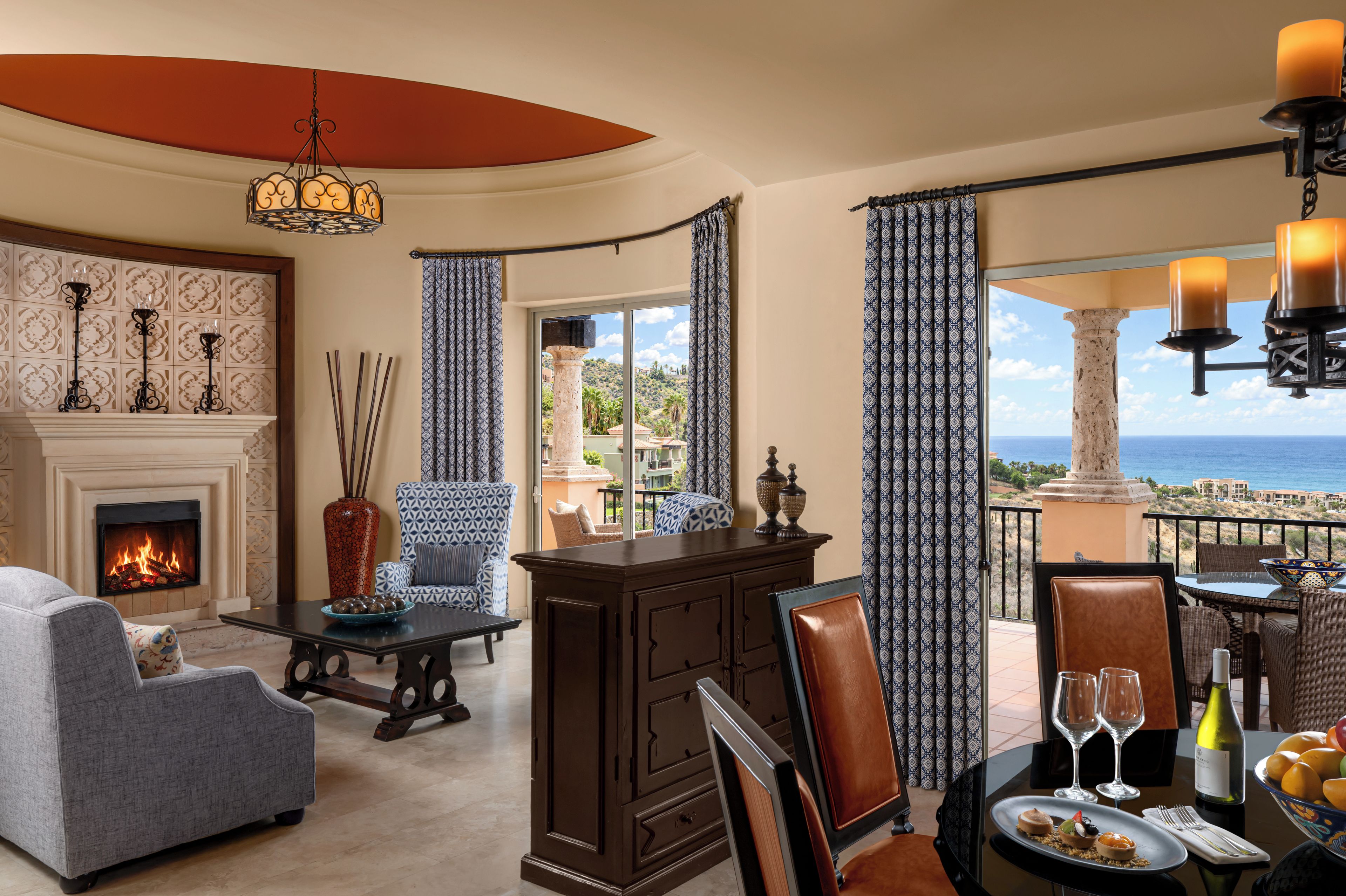A room with a fireplace, seating area, dining table with wine glasses, and a view of the sea.