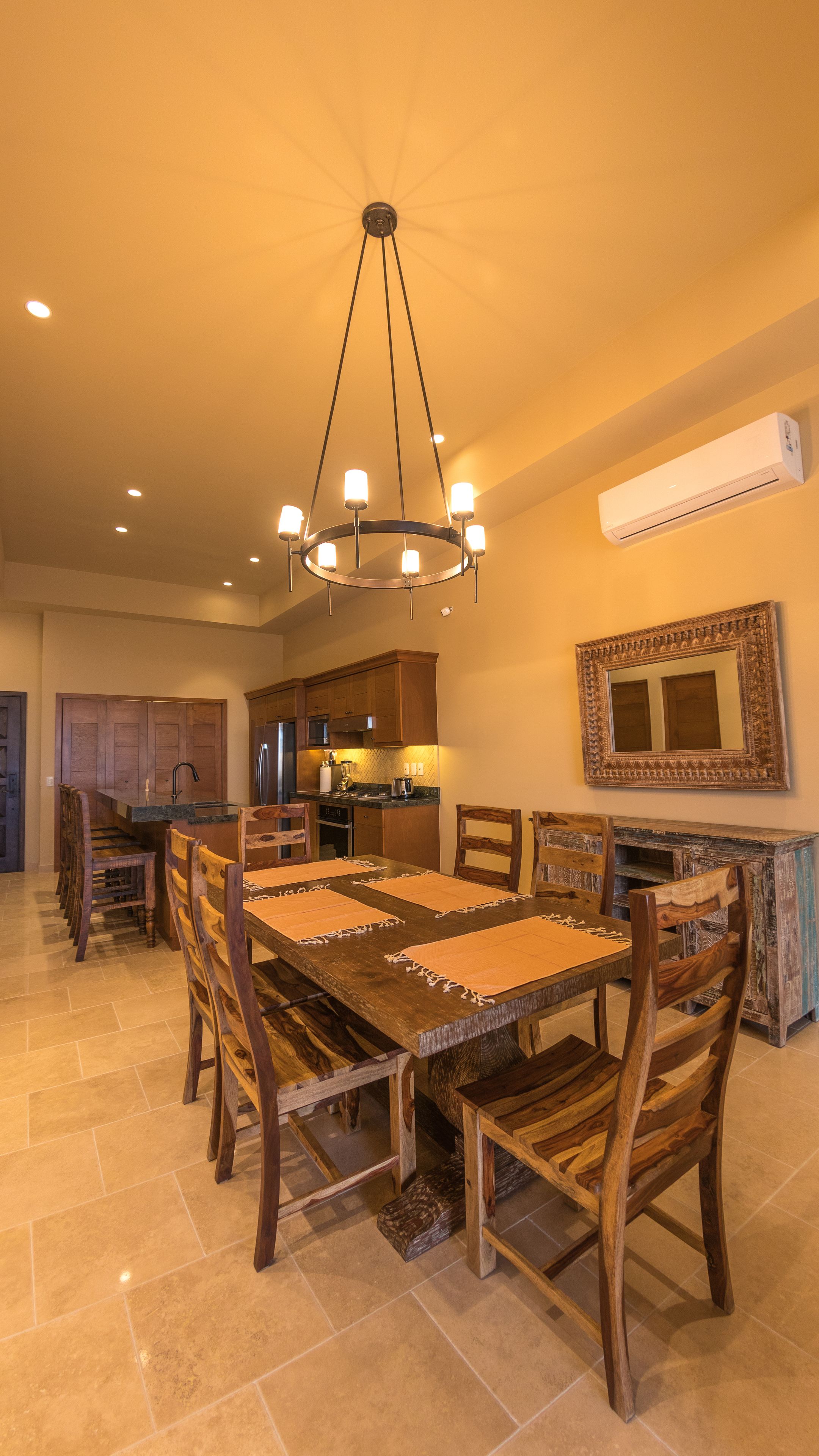 A spacious dining area with a large wooden table and chairs, a chandelier, and a kitchen in the background.