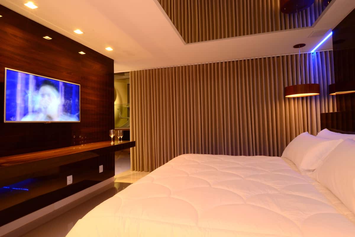 A modern hotel room with a large bed, a flat-screen TV mounted on a wooden panel, and vertical blinds on the window.