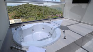 A modern bathroom with a large bathtub, a window with a view of a forested hillside, and a flat-screen TV mounted on the wall. - Le Ville (Ouro Preto)