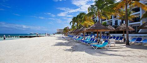 A beach with rows of blue lounge chairs and thatched umbrellas, palm trees, and a hotel in the background.