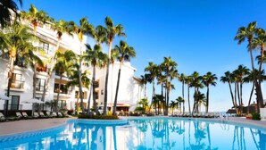 Un área de piscina con sillones de descanso y palmeras, rodeada por un edificio blanco con balcones.
