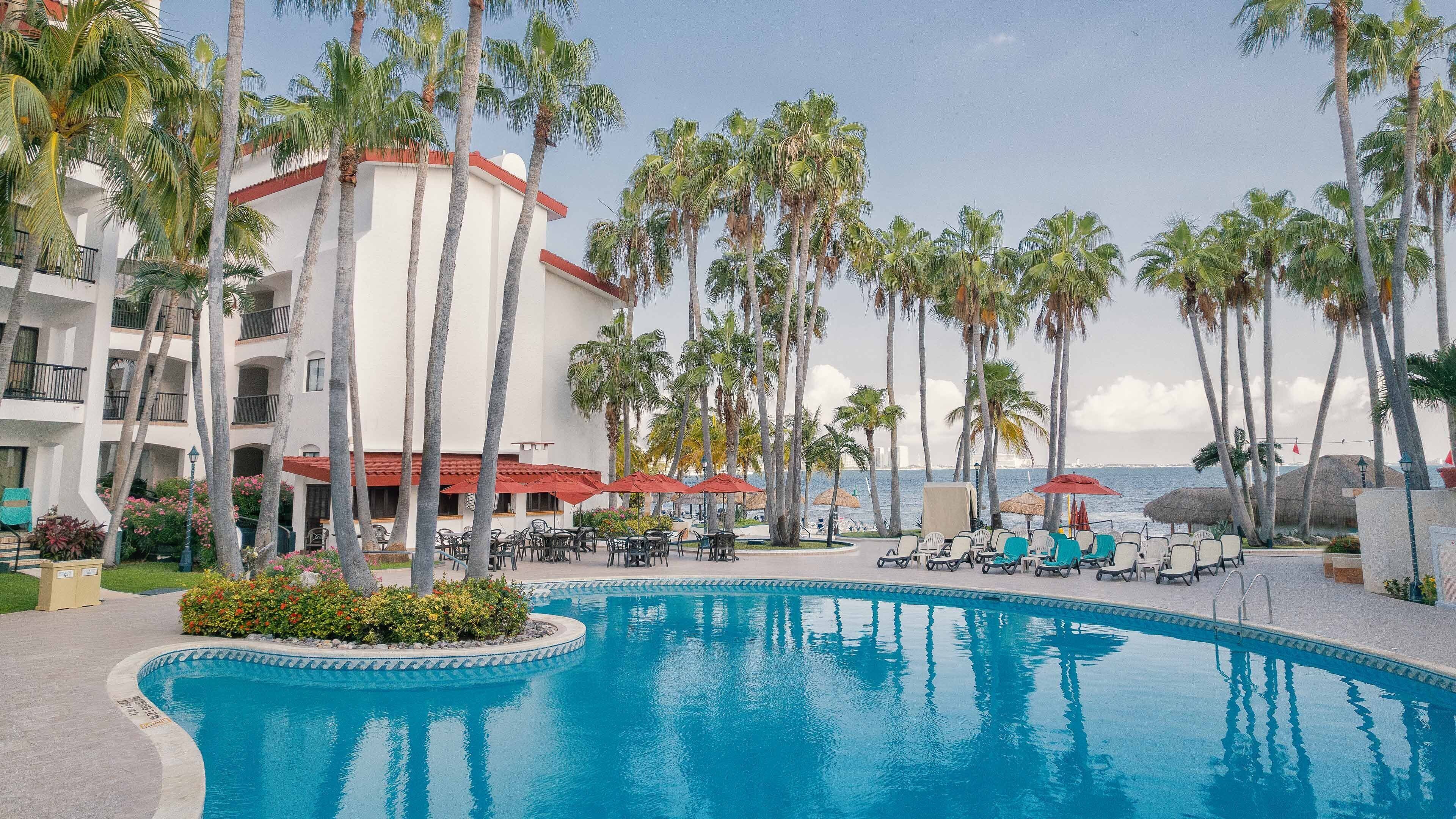 A pool surrounded by palm trees, lounge chairs, and a building with balconies.