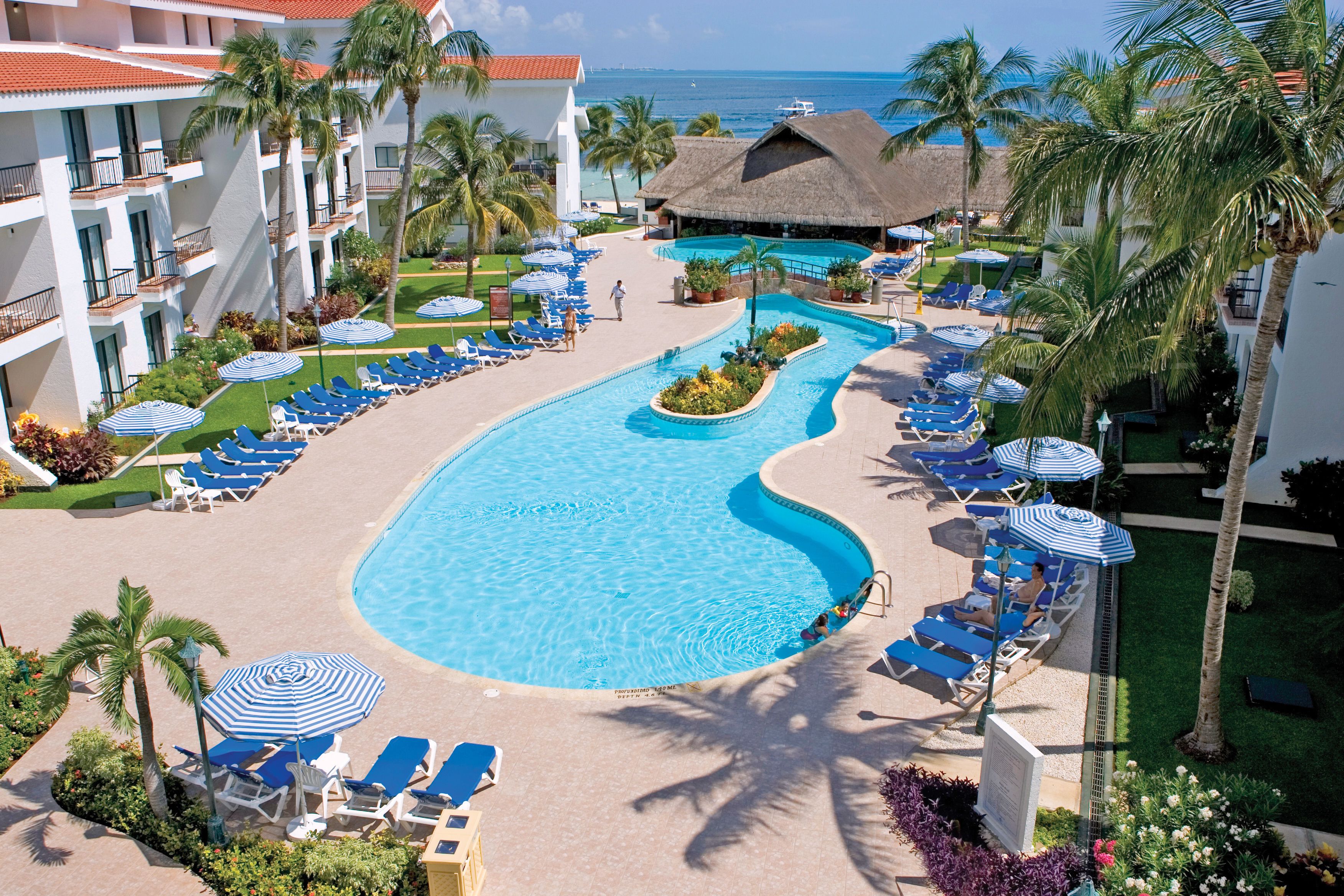 A resort pool area with lounge chairs and umbrellas, surrounded by palm trees and a view of the ocean.