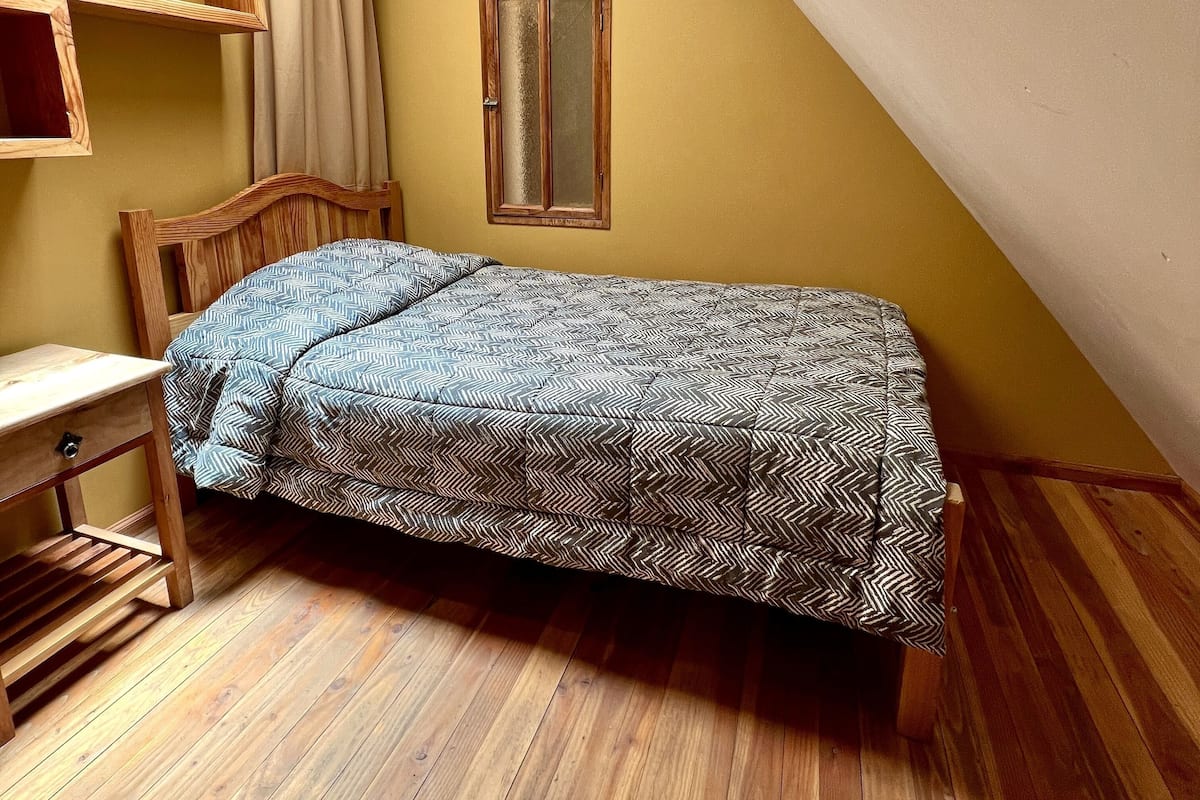 Une petite chambre avec un lit simple, un plafond en pente, un plancher en bois, un puits de lumière, une tête de lit en bois, une table de chevet et une fenêtre avec un cadre en bois.