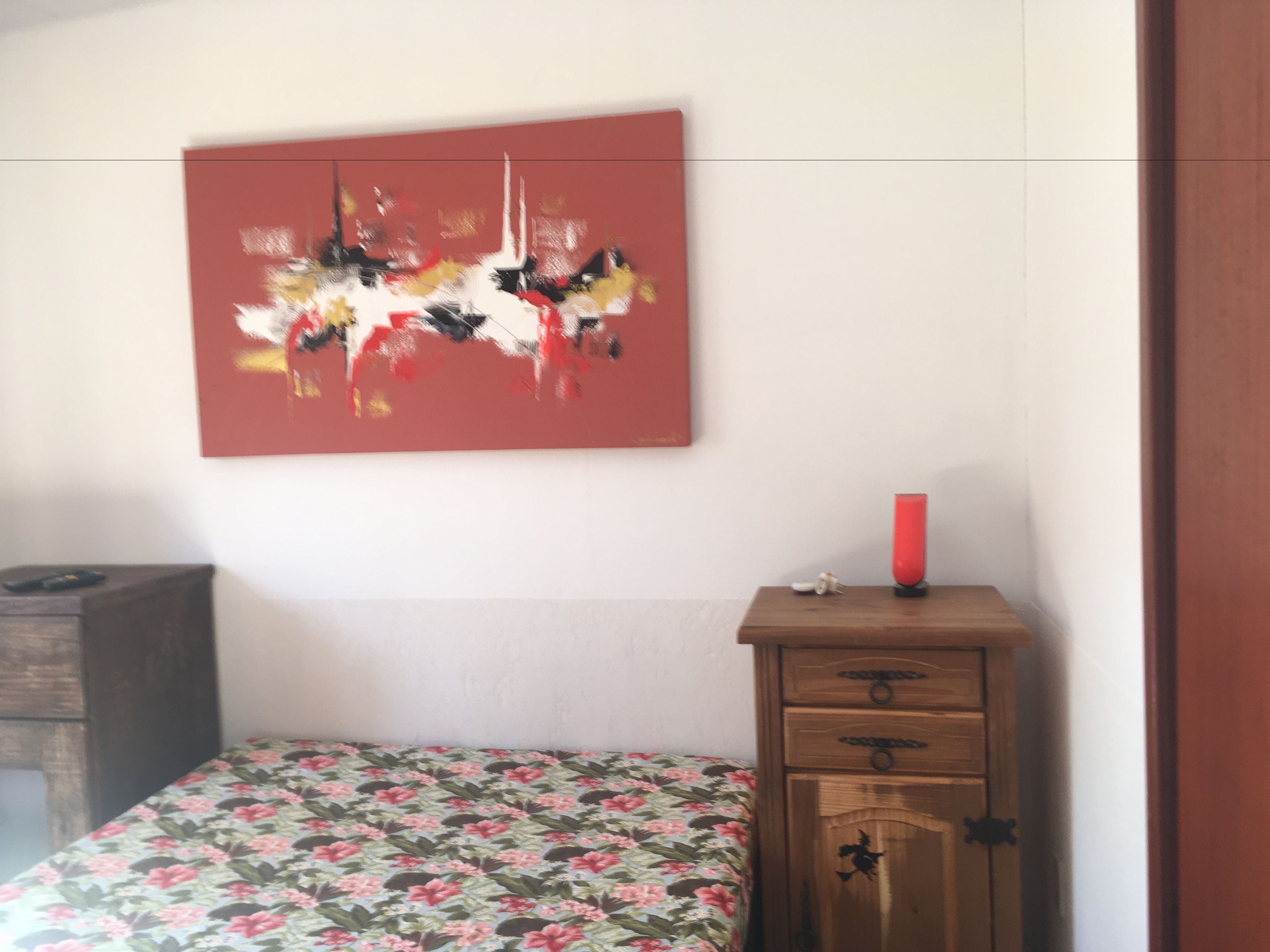 A bedroom with a floral bedspread, a wooden dresser with drawers, a red abstract painting on the wall, and a red candle on the dresser.