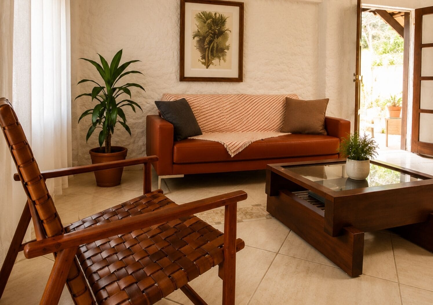 Una sala de estar con un sofá de cuero marrón, una silla tejida, una mesa de centro de vidrio, una planta en maceta e impresión enmarcadas de motivos botánicos en la pared.
