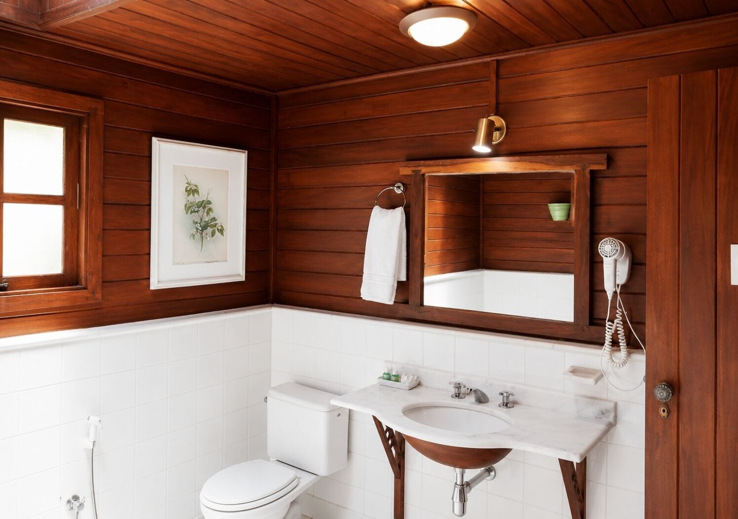 Un baño con paneles de madera, inodoro blanco, lavabo con encimera de mármol, espejo y secador de pelo en la pared.