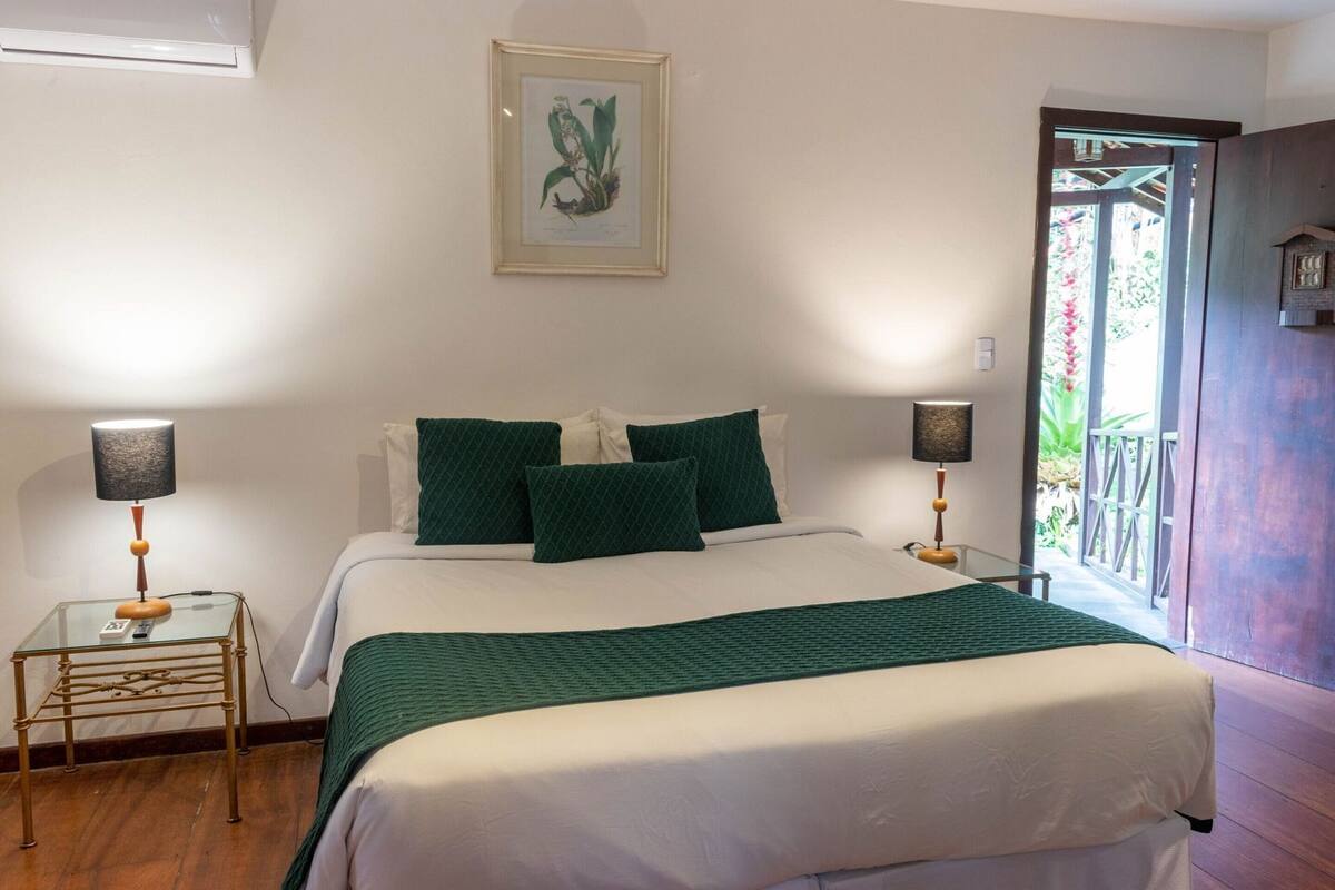 A bedroom with a bed, two bedside tables, a framed botanical illustration, and a view of the outdoors.