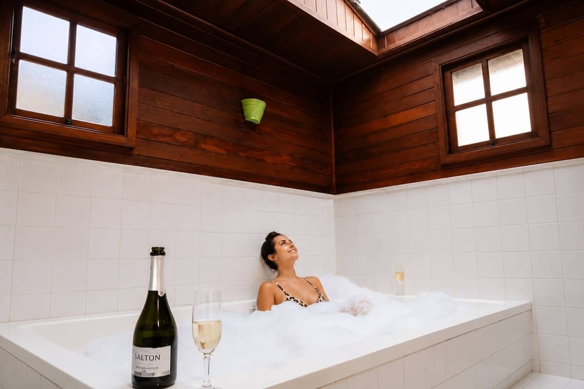 A person relaxing in a bathtub with a bottle of wine and a glass, in a room with wooden walls and windows.