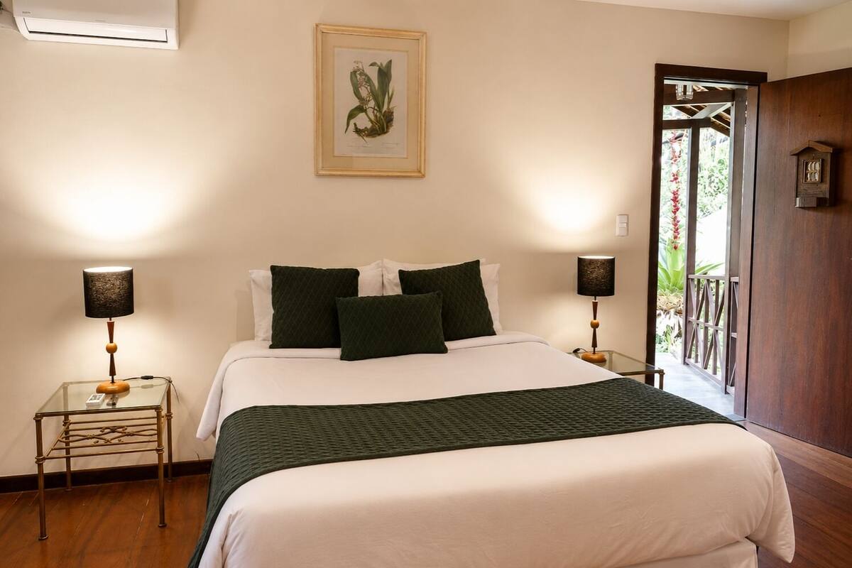 A bedroom with a bed, two bedside tables, a nightstand, a framed botanical illustration, and a wooden door leading to an outdoor area.