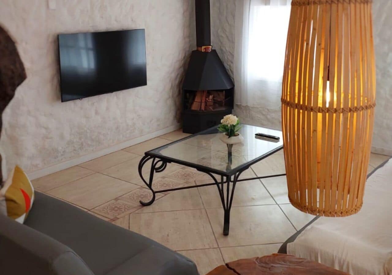 A living room with a black TV mounted on the wall, a black wood-burning stove, a glass coffee table, and a wicker lamp.