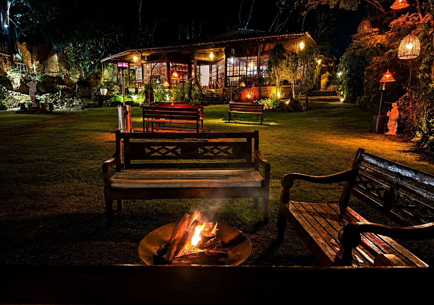 Uma área de estar ao ar livre à noite, com lareira, bancos e um edifício com janelas iluminadas.