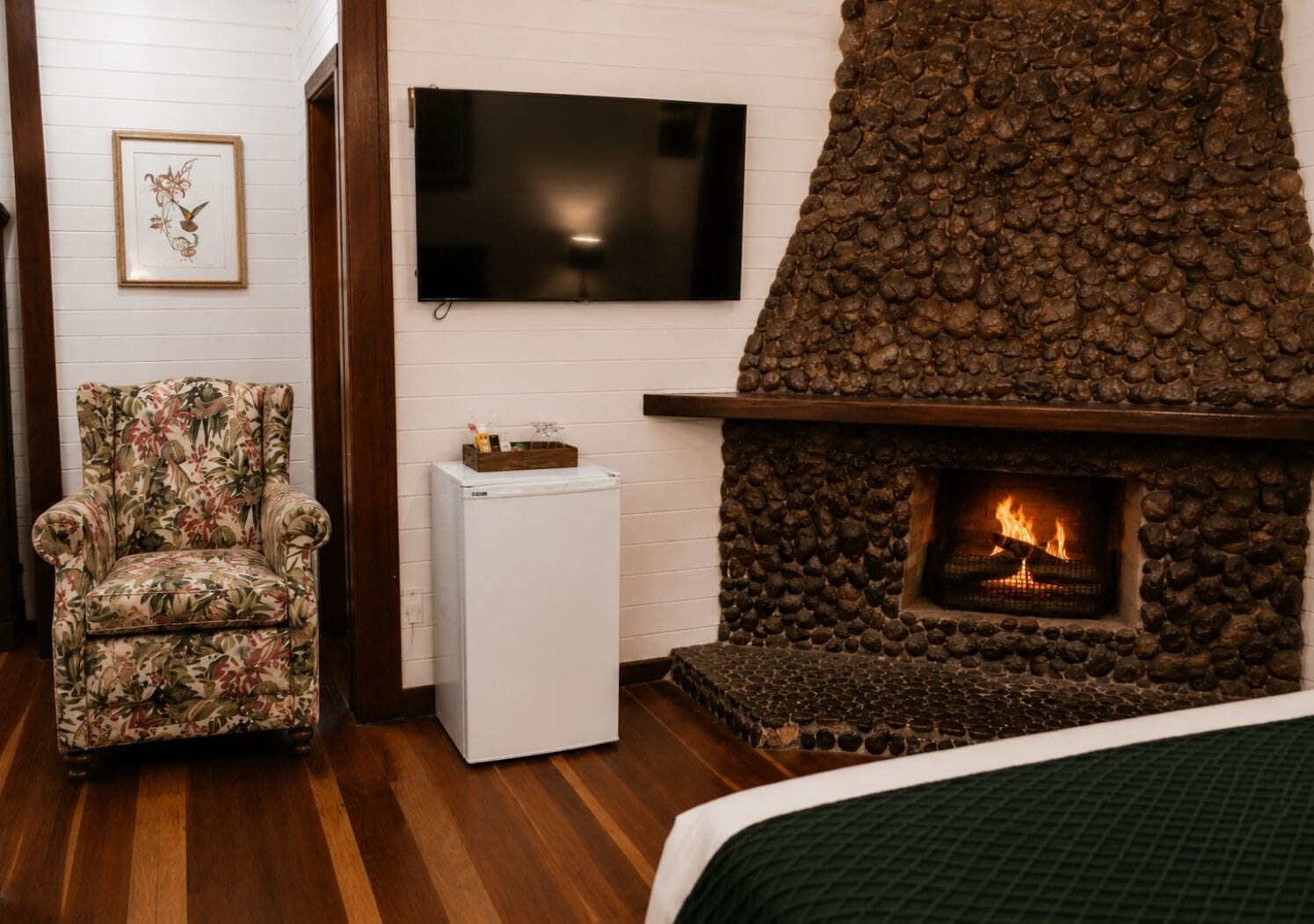 A room with a stone fireplace, a television mounted on the wall, a floral armchair, and a small white refrigerator.