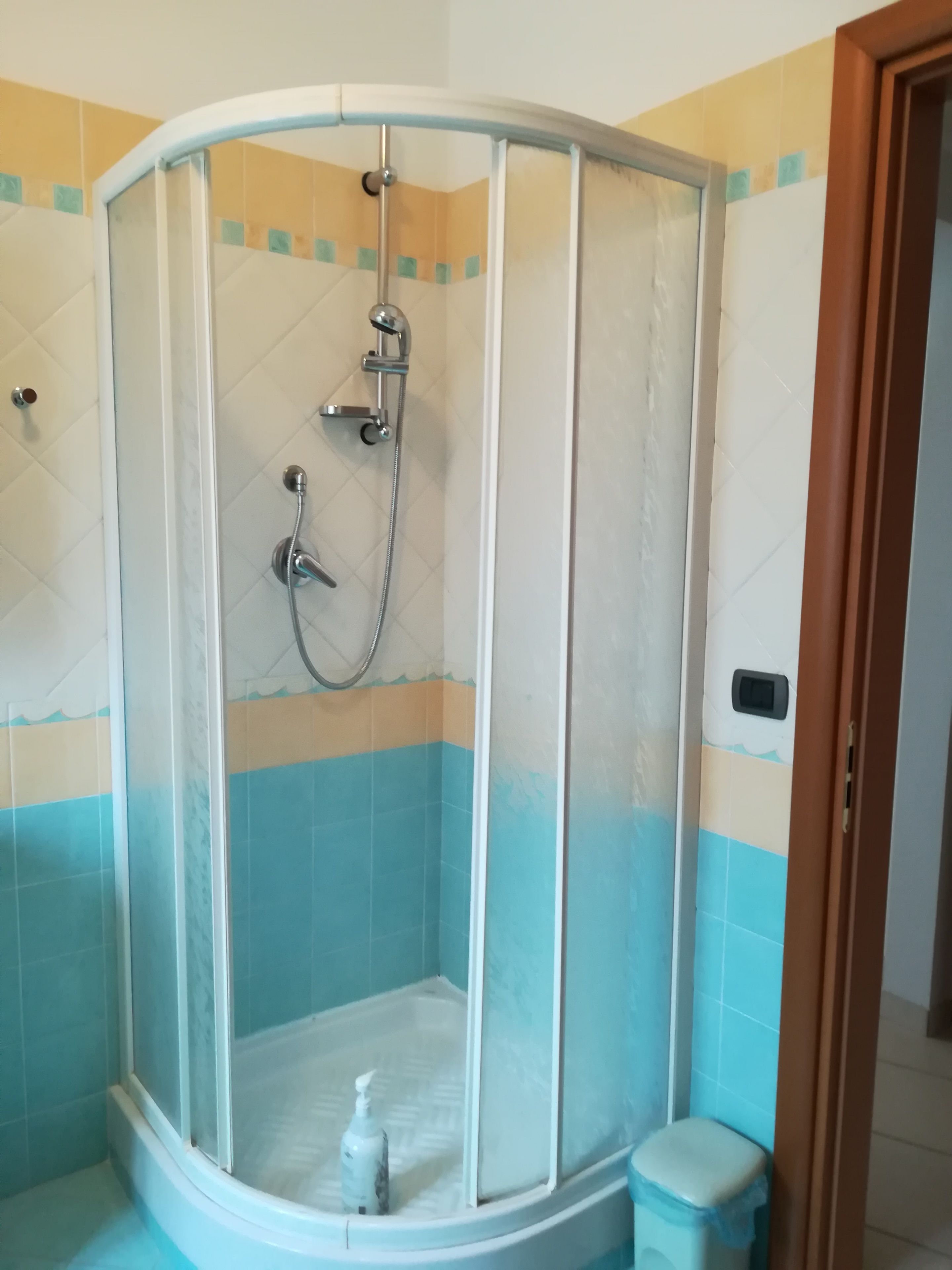 Eine Eckdusche mit Glasabtrennung, eine Handbrause und eine kleine Shampoo-Flasche auf dem Boden.