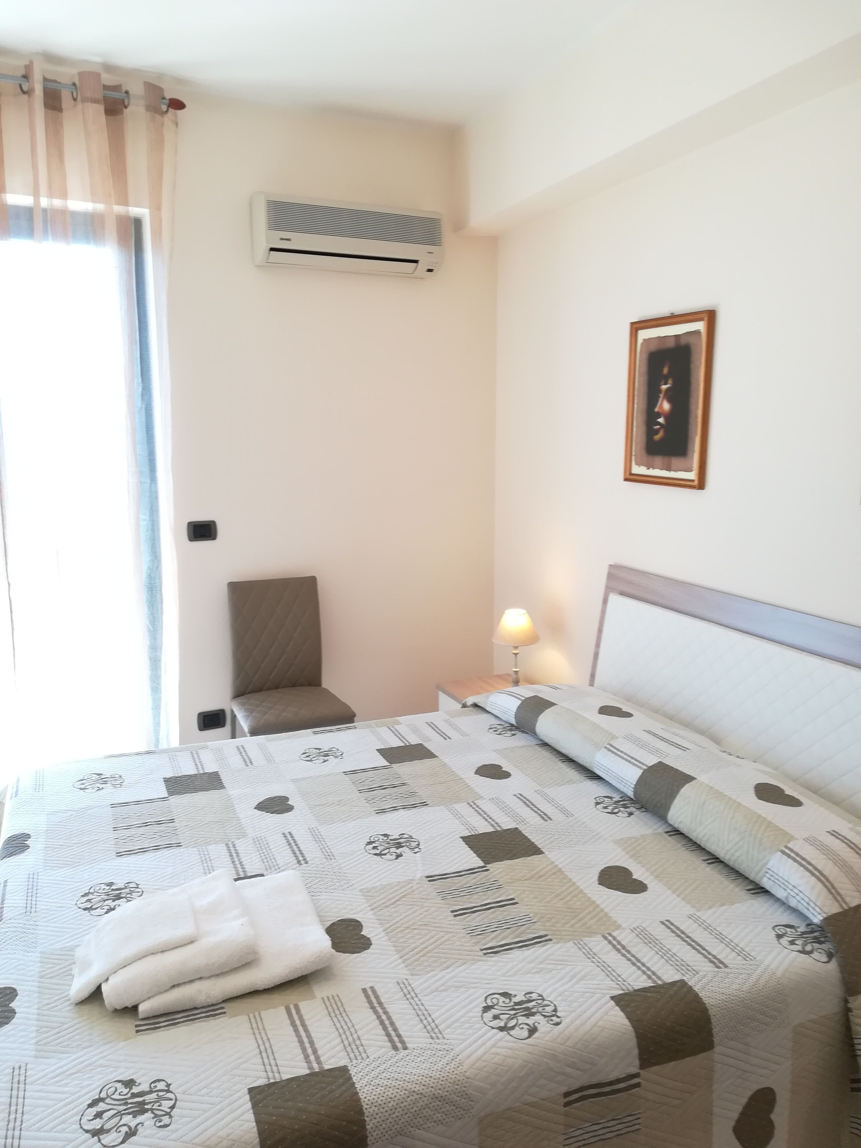 Habitación de hotel con una cama, una silla, un equipo de aire acondicionado, una lámpara y un cuadro colgado en la pared.