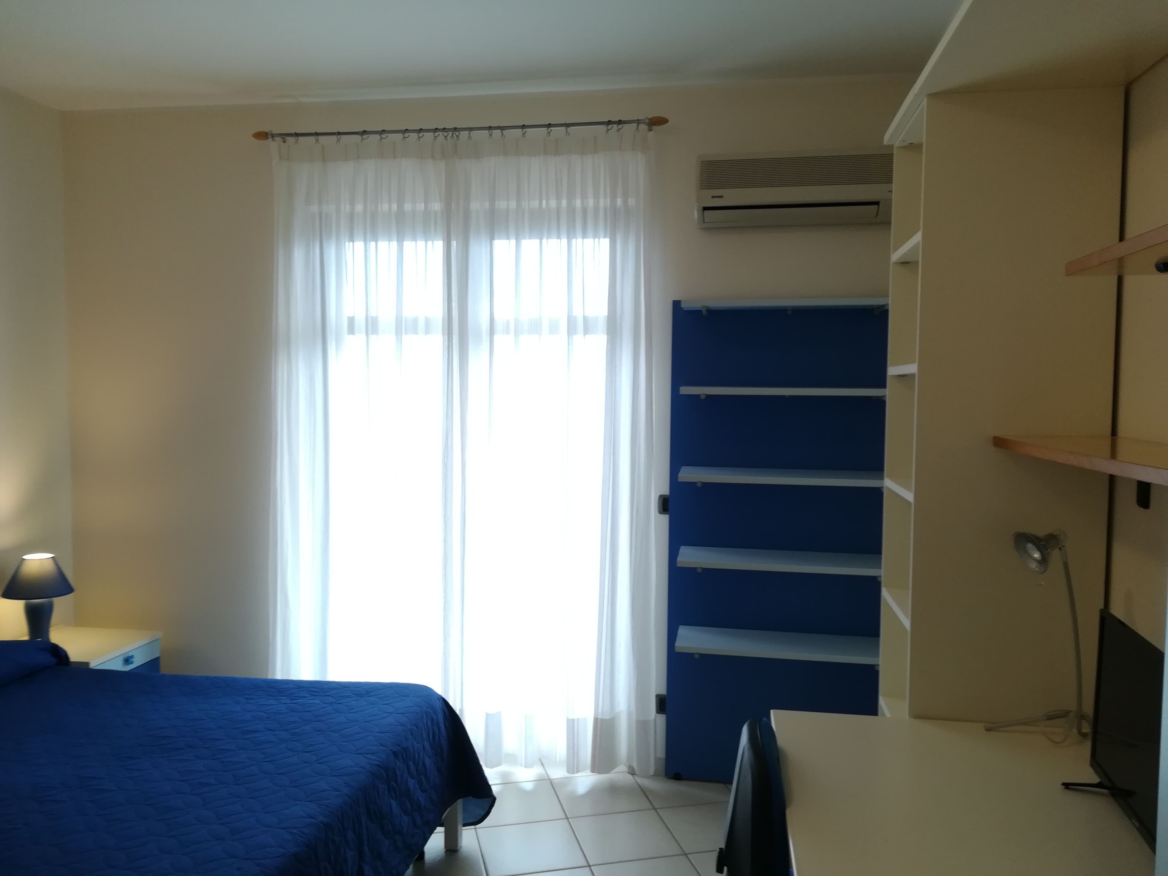 A bedroom with a bed, a desk, a chair, a lamp, a window with curtains, and a built-in shelf.