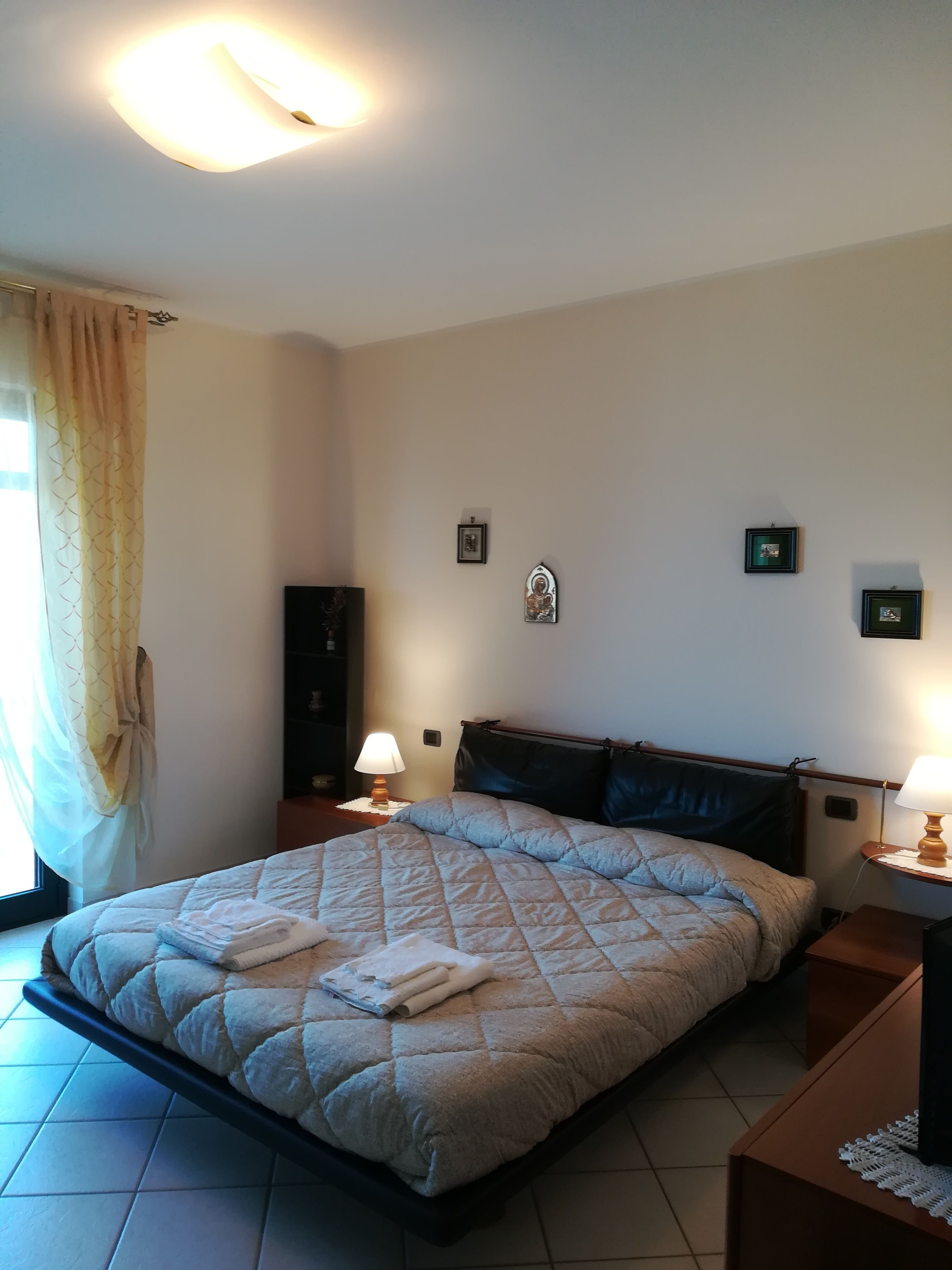Ein ordentlich bezogenes Bett mit gesteppter Decke und gefalteten Handtüchern, zwei Nachttischlampen, ein kleiner Nachttisch mit Lampe sowie ein an der Wand befestigter Bilderrahmen.