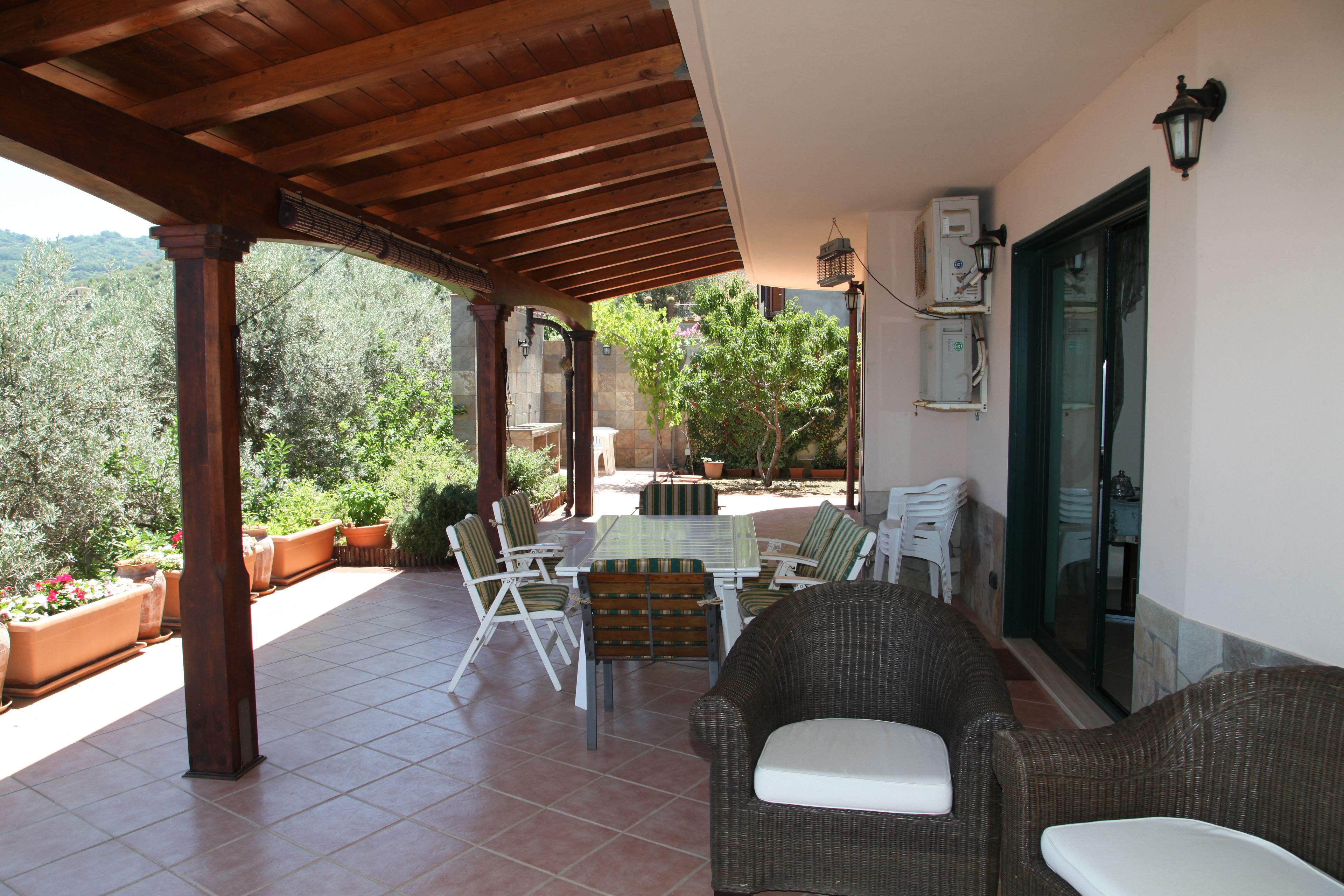An outdoor patio with a wooden pergola, a table with chairs, potted plants, and a view of the surrounding landscape.